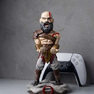 May include: A Kratos figurine standing on a circular base with a red Omega symbol. The character has a bald head with a red tattoo, a dark beard, and an open mouth. The figure wears brown leather armor, light gray pants, and dark boots, with hands positioned forward as if holding an object.