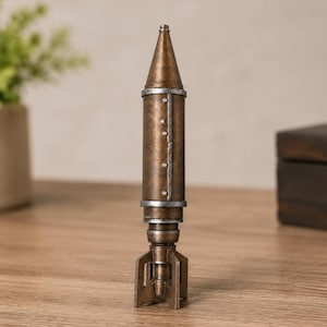 May include: A vertical, bronze-colored missile figurine with a pointed nose cone, a cylindrical body featuring a vertical seam with small silver rivets, and a four-finned tail section. The object has a weathered, metallic finish throughout.