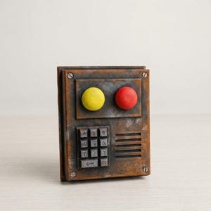 May include: A rectangular control panel with a weathered, rusted brown finish. It features one yellow circular button, one red circular button, a numeric keypad with twelve buttons, and a speaker grille. Four silver screws secure the corners.