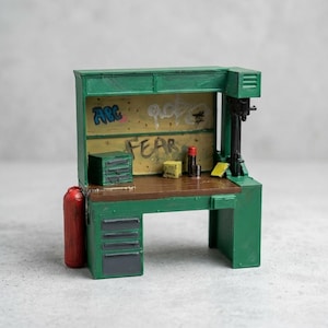 May include: A miniature green workbench featuring a brown surface, a small green tool chest, a red fire extinguisher, and a yellow backboard with graffiti text reading ABC and FEAR. The unit includes a dark gray drawer stack and a drill press.
