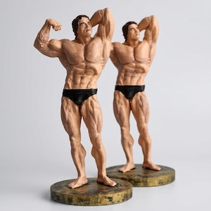May include: Two identical figurines of a muscular bodybuilder posing with one arm raised behind the head. Each figure has brown hair, wears black briefs, and stands on a circular, distressed gold-colored base with a textured, weathered finish.