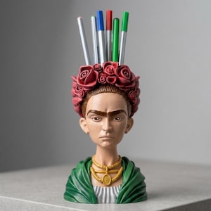 May include: A bust-shaped pencil holder depicting a person with dark hair adorned with a crown of red roses. The figure wears a green shawl over a white shirt and a gold-colored necklace. Several colored pencils are inserted into the top of the head.