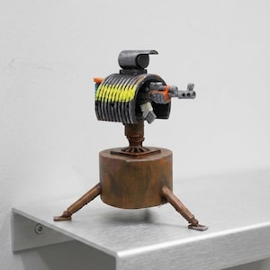 Puede incluir: Una pequeña escultura artesanal de una torreta montada sobre una base cilíndrica con tres patas. La torreta presenta una carcasa gris oscuro con una cinta de munición pintada de amarillo, un cañón naranja y una pequeña visera negra.