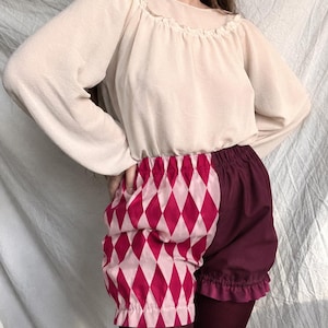 May include: A pair of high-waisted shorts featuring a split design with one side in a light pink and magenta diamond harlequin pattern and the other side in solid dark burgundy. The shorts have an elastic waistband and ruffled leg openings.