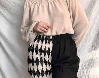 May include: A beige long-sleeved blouse with a gathered neckline and elastic cuffs paired with split-design shorts. The shorts feature a black and white diamond harlequin pattern on one side and solid black fabric on the other with ruffled hems.