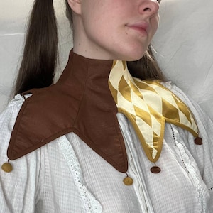 Könnte beinhalten: Ein brauner und goldener Narrenkragen mit einem Rautenmuster auf einer Seite und kleinen goldenen Pompons. Der Kragen ist aus Stoff gefertigt und wird um den Hals getragen. Der Kragen ist Teil eines Kostüms.
