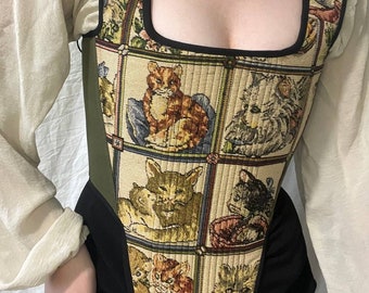 May include: A tapestry corset featuring a repeating pattern of cat illustrations in various poses. The corset has a square neckline, black trim, and a green side panel. The sleeves are long and cream-colored.