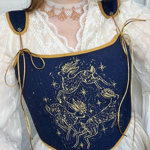 May include: A navy blue corset with a gold trim and straps. The corset features a gold illustration of deer and stars. The shoulder straps are navy blue with gold trim. The corset is worn over a white lace blouse.