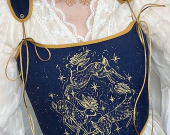 Puede incluir: Un corsé azul marino con ribete y correas doradas. El corsé presenta una ilustración dorada de ciervos y estrellas. Las correas de los hombros son azul marino con ribete dorado. El corsé se usa sobre una blusa de encaje blanco.