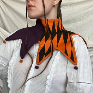 Könnte beinhalten: Ein Kragen im Narren-Stil mit geteiltem Design, wobei eine Seite in einfarbigem Dunkellila und die andere Seite in einem orange-schwarzen Rautenmuster gehalten ist. Der Kragen hat spitze Kanten, die jeweils mit einem kleinen runden Bommel enden.