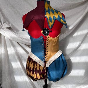 Op de afbeelding: Een kleurrijk narrenkostuum uitgestald op een mannequin. Het kostuum heeft een tweekleurig lijfje, met blauwe en bruine delen, en een bijpassende pofbroek. De kraag van de nar is een combinatie van bordeauxrode en gele ruitpatronen, met blauwe pompons.