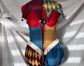 May include: A colorful jester costume displayed on a mannequin. The costume features a two-tone bodice, with blue and tan sections, and a matching pair of bloomers. The jester's collar is a combination of burgundy and yellow diamond patterns, with blue pom-poms.