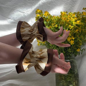 May include: A pair of wrist cuffs featuring a gathered, cream-colored fabric with a repeating brown diamond pattern. Each cuff is finished with solid dark brown fabric borders at the top and bottom edges, creating a structured, ruffled appearance.