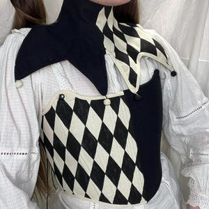 Könnte beinhalten: Ein schwarz-weißes Harlekin-Stil-Mieder- und Kragenset. Das Mieder hat ein Rautenmuster, während der Kragen ein Schachbrettmuster aufweist. Das Set enthält kleine, dekorative Pompons. Das Kleidungsstück wird über einer weißen Bluse getragen.