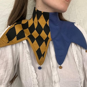 Könnte beinhalten: Ein Narrenkragen mit hohem Hals und Schulterdrappe. Der Kragen hat ein Harlekinmuster in Schwarz und Gold mit einem durchgehenden blauen Abschnitt. Kleine, dekorative blaue und braune Pompons hängen von den Spitzen.