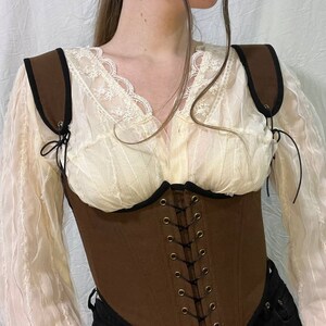 May include: A brown canvas corset with black trim and adjustable straps. The corset has a lace-up front with brass eyelets and is worn over a white lace blouse.