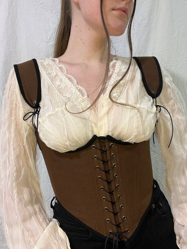 May include: A brown canvas corset with black trim and adjustable straps. The corset has a lace-up front with brass eyelets and is worn over a white lace blouse.