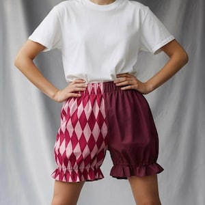 Peut inclure: Un t-shirt blanc à col rond associé à un short taille haute. Le short présente un design divisé : un côté avec un motif de losanges roses et rouges, l'autre bordeaux uni. Le short a des ourlets à volants.