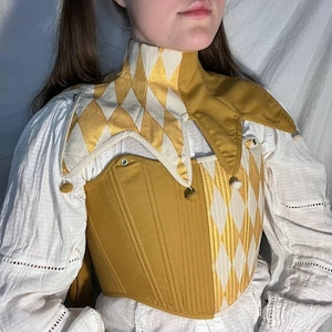 Könnte beinhalten: Senfgelbes Korsett mit hohem Kragen und Rautenmuster in Weiß und Gold. Das Korsett hat ein strukturiertes Mieder mit vertikalen Nähten und kleinen dekorativen Knöpfen. Der Kragen hat ein Narren-Design.
