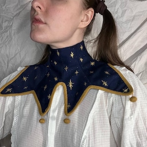 Puede incluir: Un cuello azul marino con bordado de estrellas doradas y ribete dorado. El cuello tiene un borde dentado y dos pequeños pompones dorados. Diseñado para llevar alrededor del cuello.