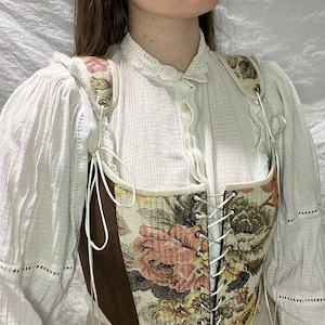 May include: A brown and white floral corset with a lace-up front and adjustable straps. The corset is made of a woven fabric with a floral pattern and has a brown leather trim.