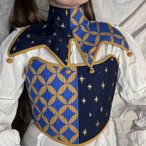 Könnte beinhalten: Ein blau-goldenes, gestepptes Mieder mit hohem Kragen und Schulterteilen im Narrenstil. Das Mieder hat auf einer Seite ein geometrisches Muster und auf der anderen Seite ein Sternennacht-Design. Goldbesatz und kleine Bommel.
