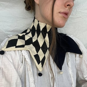 May include: A black and white checkered fabric collar with a pointed design. The collar has a black fabric trim and white pom-poms on the ends.