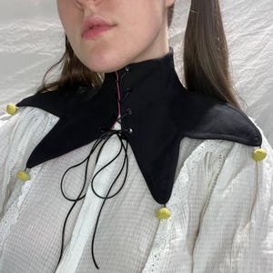 May include: A black velvet collar with a pointed, jester-like design. The collar has a pink lining and is laced up the front with black cord. Yellow pom-poms are attached to the points. The collar is worn over a white blouse.