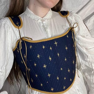 Pode incluir: Um espartilho azul marinho com detalhes de estrelas e salpicos dourados. O espartilho tem acabamentos e alças douradas. A peça é amarrada com fio dourado. O espartilho é usado sobre uma blusa branca com gola de renda.