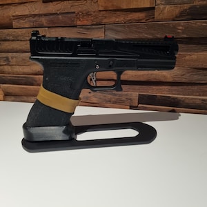 Könnte beinhalten: Eine schwarze Pistole mit einem braunen Griff wird auf einem schwarzen Ständer präsentiert. Die Waffe hat ein rotes Visier und einen silbernen Abzug.