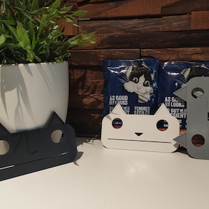 Puede incluir: Tres soportes con forma de gato para bolsas de comida para gatos. Los soportes son de plástico blanco, gris y negro. Las bolsas son azules y blancas con el texto "As good as it looks" y "Tendres Effiles".