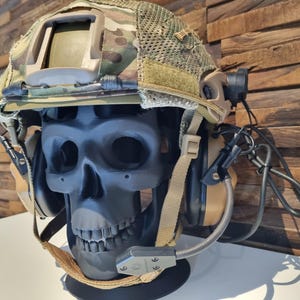 Può includere: Un casco mimetico con un supporto a forma di teschio nero e un auricolare color beige con un microfono attaccato.