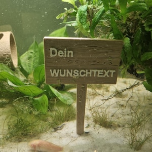 Könnte beinhalten: Ein kleines Holzschild mit dem Text "Dein WUNSCHTEXT" in hellbrauner Schrift. Das Schild ist auf einem Stock befestigt und von grünen Pflanzen und weißem Sand umgeben.