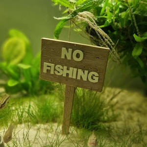 Könnte beinhalten: Ein Miniatur-Holzschild mit dem Text "NO FISHING" in weißen Buchstaben, platziert in einem Aquarium mit grünen Pflanzen und Sand.