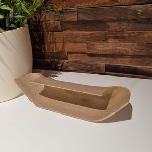 Könnte beinhalten: Eine beige, gebogene Ablage mit einem erhöhten Regal. Die Ablage besteht aus einem strukturierten Material und steht auf einer weißen Oberfläche. Ein weißer Keramiktopf mit grünen Pflanzen befindet sich im Hintergrund, zusammen mit einer Holzwand.
