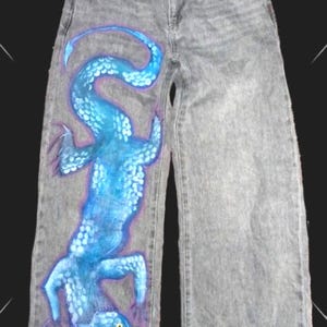 Peut inclure: Jean gris avec un grand dragon bleu peint sur le devant. Le dragon est détaillé avec des écailles et souligné de bleu foncé. Le jean a une conception classique à cinq poches. Le jean mesure environ 100 cm de long.