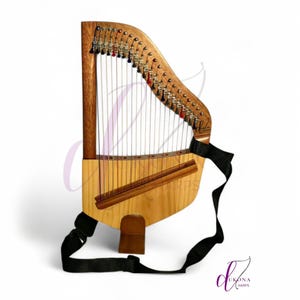 May include: A wooden lap harp with a light-colored soundboard and a darker wood frame. The instrument features numerous vertical strings, metal tuning pins with black and red accents, and a black adjustable nylon strap for carrying or playing.
