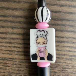 May include: A black pen with a decorative top. The top features a black and white striped bead, a pink bead, and a white rectangular charm with a woman holding a pink cup. The word "BOSS" is visible on the cup.