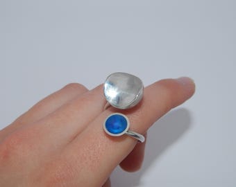 May include: Two silver rings. One ring has a large, irregular, silver-colored top. The other ring has a small, round, blue top. Both rings are on a finger.