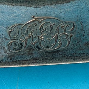 May include: A metallic surface with a weathered, blue-toned patina featuring an intricate, hand-engraved monogram of the letters F, E, and B. The letters are interconnected with ornate, swirling flourishes and fine lines etched into the metal.