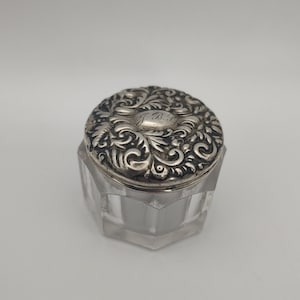 Puede incluir: Un pequeño frasco de vidrio transparente con una tapa de plata. La tapa está decorada con un intrincado diseño floral y tiene una inscripción que dice "J.B."