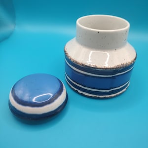 Könnte beinhalten: Ein Keramiktopf mit Deckel, mit einer weiß gesprenkelten Oberseite und einem blau-weiß gestreiften Körper. Der Deckel ist rund und passt zum Farbschema des Topfes. Der Topf ist etwa 10 cm hoch.