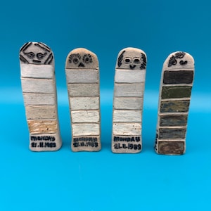 May include: Four ceramic sculptures with faces and layered bodies, each with a different expression. The sculptures are in shades of white, brown, and gray, with blue text at the base indicating the day and date.