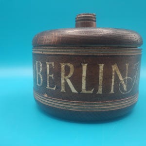 May include: A round, dark brown wooden container with a lid and a small handle on top. The word "BERLIN" is inscribed in a light colour around the container's body. The container sits against a blue background.