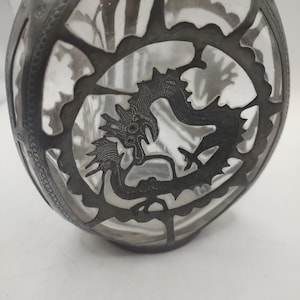May include: A clear glass bottle with a silver metal design featuring a dragon. The bottle has a textured surface and a round shape.