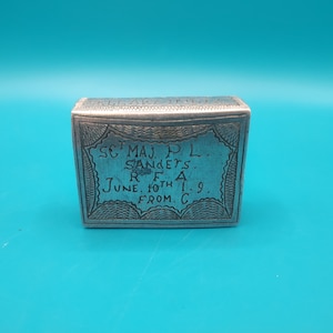 Puede incluir: Pequeña caja rectangular plateada con texto grabado y bordes decorativos. El texto incluye "SC MAJ P.L. SANDERS. R.F.A. JUNE. 10TH 1.9. FROM. C." La caja está sobre un fondo verde azulado.