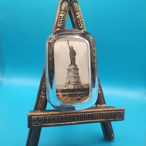 Puede incluir: Un marco de fotos decorativo estilo caballete con una parte frontal de vidrio transparente. El marco contiene una imagen en blanco y negro de la Estatua de la Libertad. El marco está sostenido por un caballete de madera de color marrón y dorado.