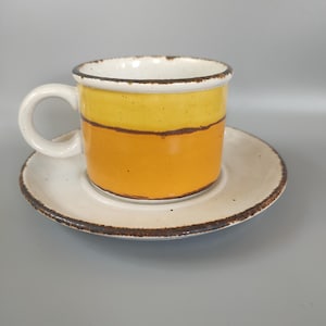 Könnte beinhalten: Ein Keramik-Kaffeebecher und Untertassen-Set. Der Becher ist zylindrisch mit einem weißen Henkel und ist in zwei Abschnitte unterteilt: gelb und orange. Die Untertasse ist weiß mit einem braunen Rand. Das Set ist für Heißgetränke konzipiert.
