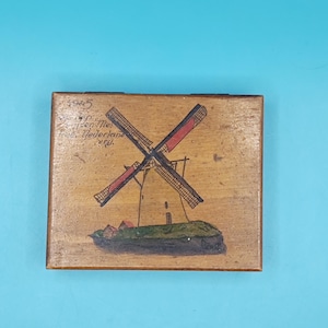 Puede incluir: Caja de madera con una escena de molino de viento pintada a mano. El molino tiene aspas rojas y negras, sobre un fondo marrón claro. La caja tiene texto en la esquina superior izquierda, y el molino está sobre una colina verde.