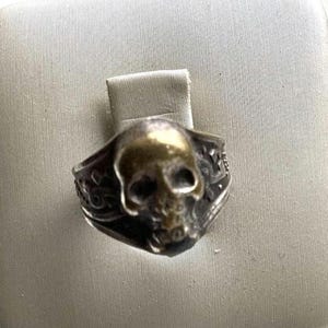 Puede incluir: Anillo plateado con diseño de calavera y rostro dorado. El anillo presenta detalles intrincados alrededor de la banda, con un acabado oscuro y oxidado. Los rasgos de la calavera están claramente definidos.